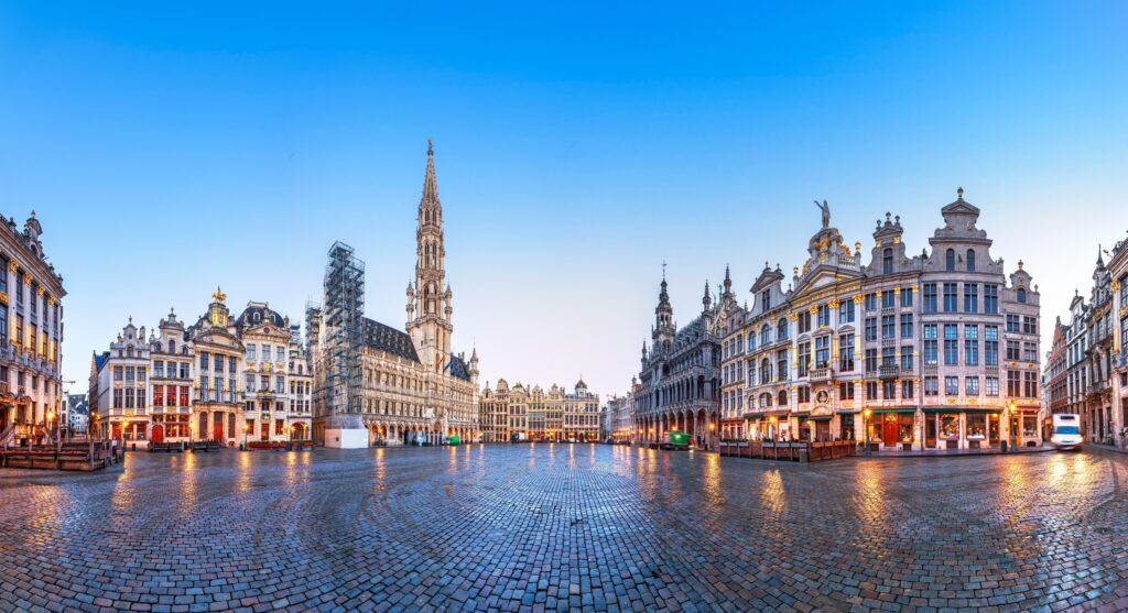 Monteurunterkünfte in Belgien mieten: Eine weitläufige Panoramaaufnahme des historischen Grand Place in Brüssel mit seinen prachtvollen Gildehäusern und dem gotischen Rathausturm unter einem klaren blauen Abendhimmel.