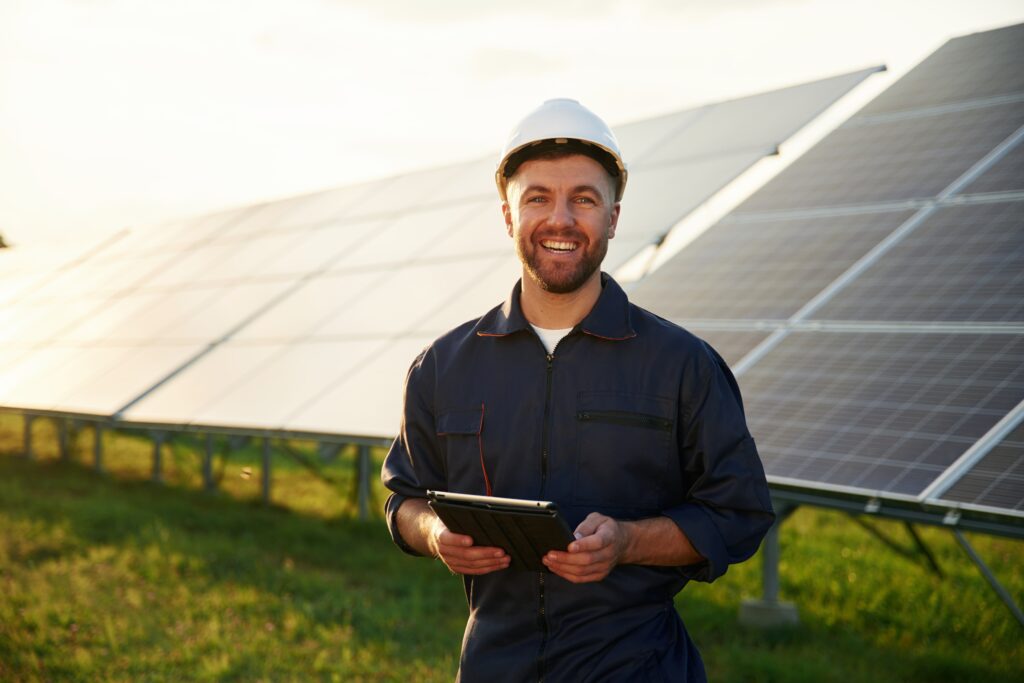 Handwerkerwohnungen: Ein lächelnder Techniker in blauer Arbeitskleidung und weißem Schutzhelm steht mit einem Tablet vor einer großflächigen Photovoltaikanlage im Sonnenlicht.