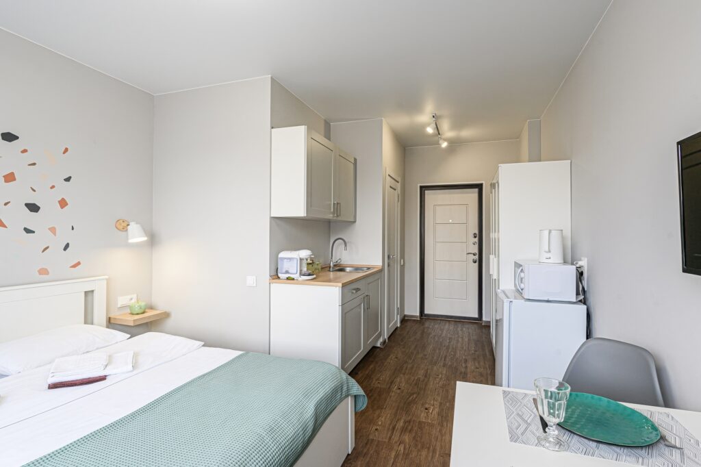 Eine helle, moderne eingerichtete Monteurwohnung mit einem weißen Bett im Vordergrund, einer angrenzenden grauen Küchenzeile mit Holz-Arbeitsplatte und einem kleinen Essbereich. Der Raum besticht durch ein minimalistisches Design mit sanften Farben und funktionaler Aufteilung.