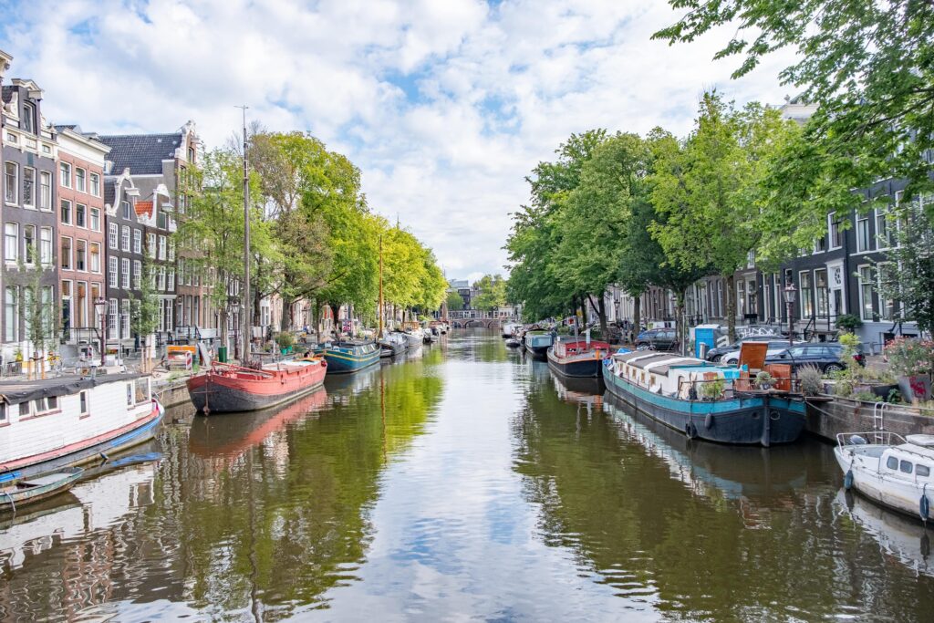Monteurunterkünfte in den Niederlanden mieten: Eine idyllische Gracht in Amsterdam mit Hausbooten an beiden Ufern, gesäumt von historischen Gebäuden und grünen Bäumen unter leicht bewölktem Himmel.